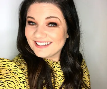 Smiling woman with long dark hair wearing a yellow patterned top, looking at the camera against a plain background.