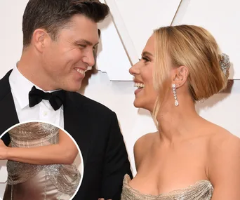 A couple in formal attire smiling at each other on a red carpet, with a close-up of an arm inserted.