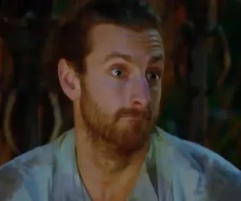 A person with long hair and a beard looks surprised on a reality TV show set with blurred background.