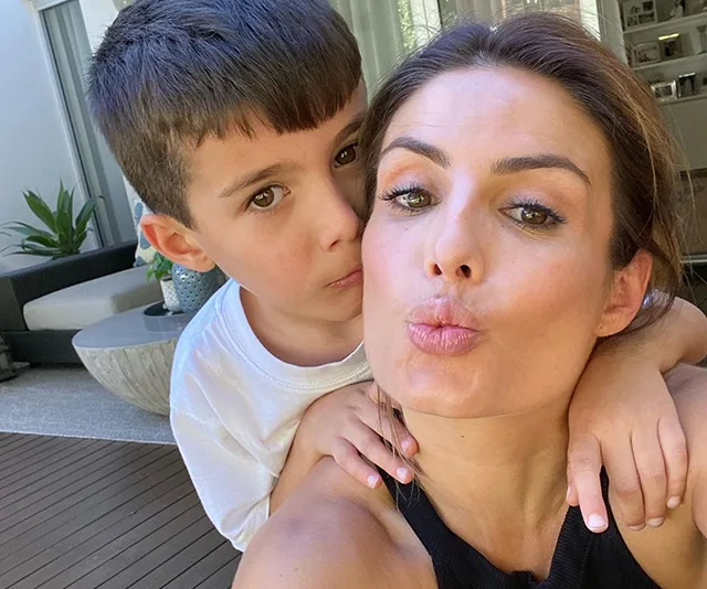 A woman takes a playful selfie with a young boy embracing her, both with relaxed expressions in a cozy outdoor setting.