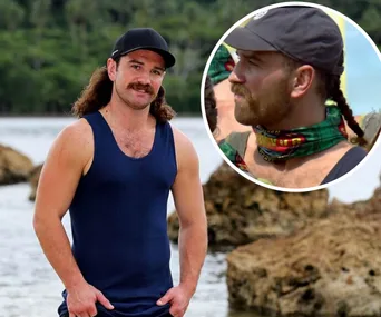 John Eastoe from Survivor All Stars 2020, wearing a blue tank and a cap, standing by rocks near the water.