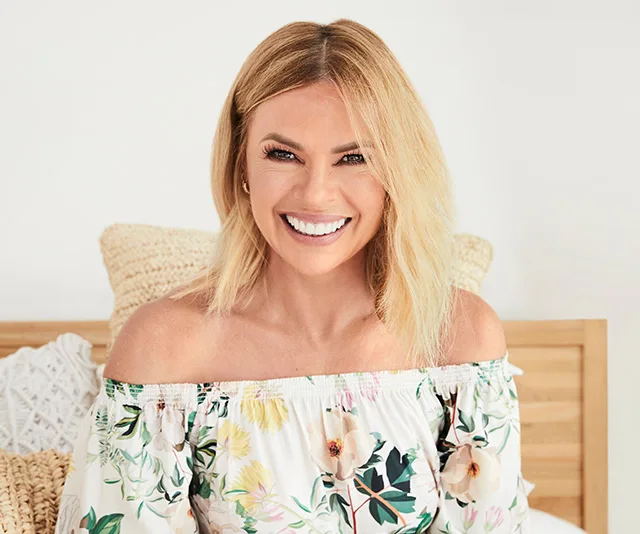 Smiling woman with blond hair in floral off-shoulder top, sitting on a bed with light wood and textured cushions.