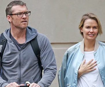 Two individuals walking outdoors, one with glasses and a backpack, the other in a light blue jacket.