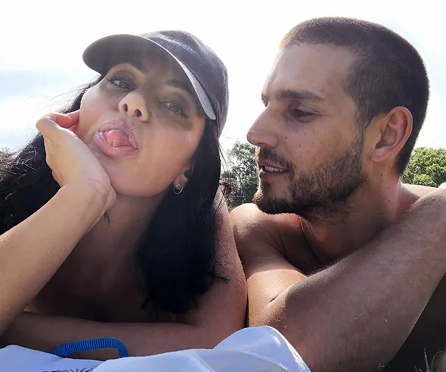 Couple relaxing outdoors; woman playfully sticks out tongue, man looks at her.