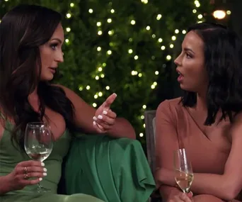 Two women sit and converse intensely, holding wine glasses, with a background of twinkling lights.