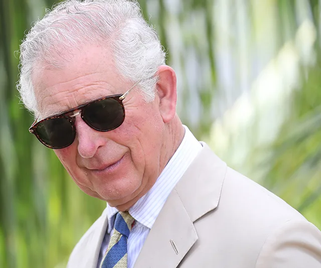 Senior man in sunglasses and light suit outdoors, head tilted slightly, smiling gently against a blurred leafy background.