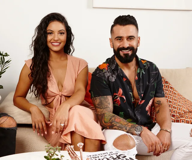 Couple seated on a couch, smiling. The woman wears a peach dress, and the man sports a floral shirt with tattoos.
