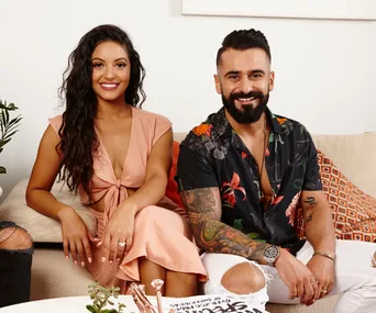 Couple seated on a couch, smiling. The woman wears a peach dress, and the man sports a floral shirt with tattoos.