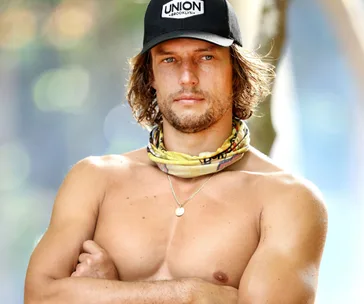 Shirtless man with a beard, wearing a black cap and necklace, arms crossed, with blurred background.