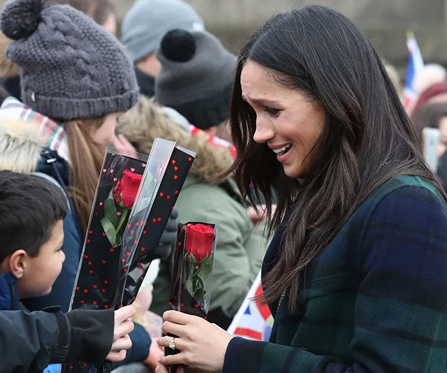 Meghan Markle smiles as she receives a rose from a child in a crowd, surrounded by onlookers.