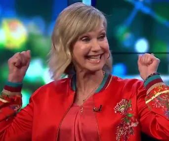 Olivia Newton-John smiling and cheering in a colorful jacket on a TV show set.