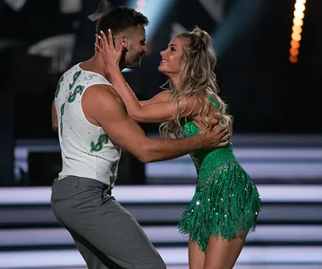 A couple gracefully dances on stage; the woman in a green sparkly dress and the man in a white tank top with decorative patterns.