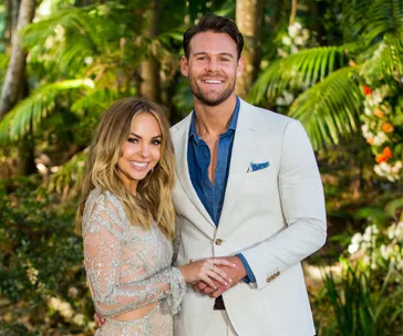 A couple smiling in a lush garden setting, with the woman in a beaded dress and the man in a light suit.
