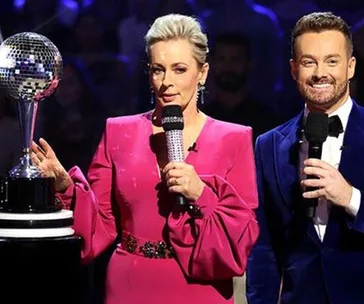 Two hosts on stage with microphones, one in a pink dress, near a mirrorball trophy, likely from a dance show.