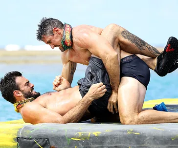 Two men wrestle playfully on a floating platform during a Survivor challenge in sunny, clear weather.