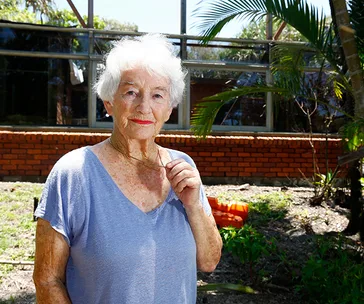 Elderly woman with white hair stands outside a brick building, holding her glasses with a calm expression.