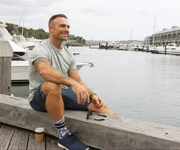 Man sitting on a wooden beam by the marina, smiling with boats and water in the background.