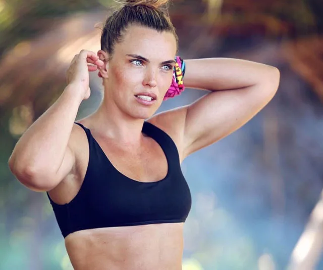 A woman in a black sports bra outdoors, adjusting her hair, with colorful bracelets on her wrist.