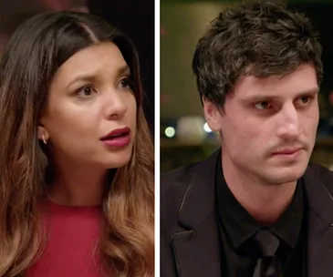 Two individuals at a dinner table; a woman with long hair looks surprised, and a man in a suit listens intently.