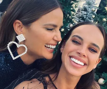 Two women smiling joyfully in front of decorated Christmas trees.