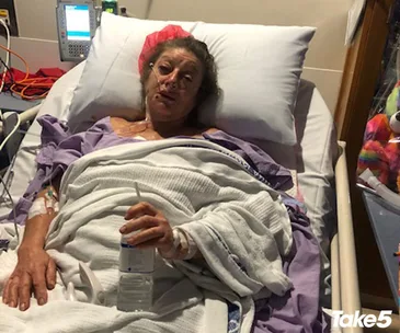 Elderly woman in hospital bed, wearing oxygen and holding a water bottle, with medical equipment in the background.