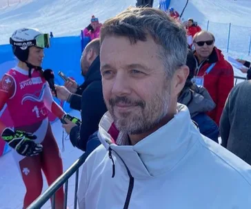 Man in white jacket smiling at a ski event; skier and media in background.