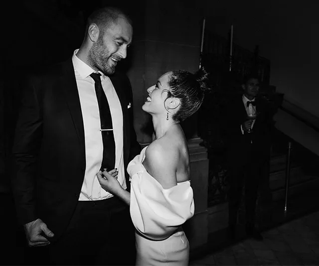 A couple smiling at each other in formal attire, a man in a suit and a woman in an off-shoulder dress, at an elegant event.