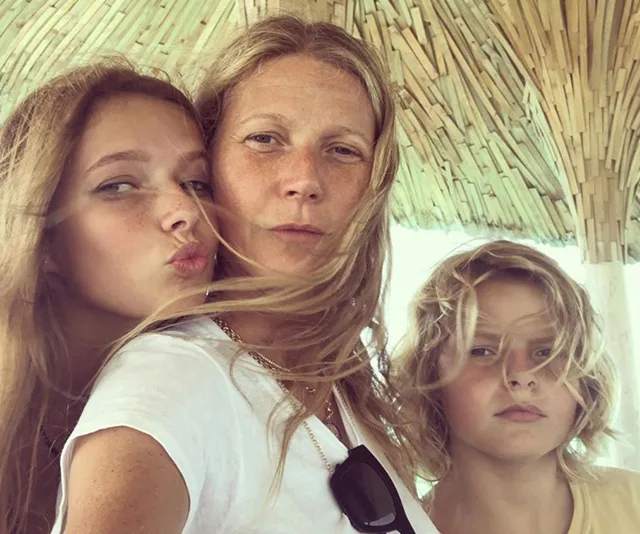 Family selfie under a thatched roof, with two children and an adult, with wind blowing their hair.