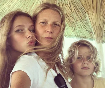 Family selfie under a thatched roof, with two children and an adult, with wind blowing their hair.