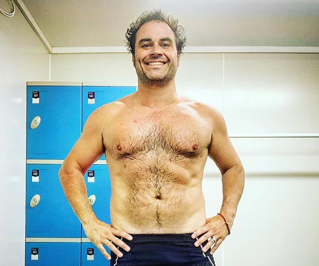 Shirtless man smiling in front of blue lockers in a gym setting, showcasing weight loss transformation.