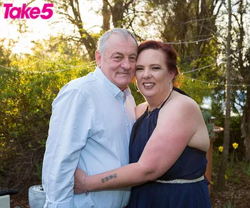 An elderly couple smiling, embracing in a garden setting with fairy lights during sunset.