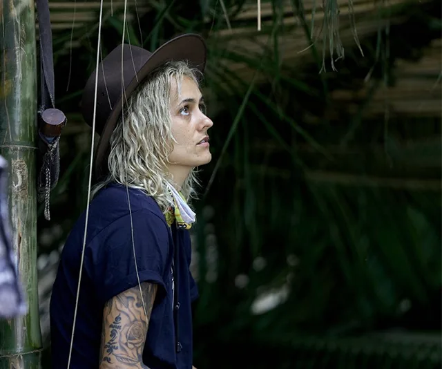 A person with a hat and tattoos sits thoughtfully in a jungle setting, surrounded by greenery.