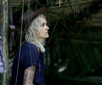 A person with a hat and tattoos sits thoughtfully in a jungle setting, surrounded by greenery.