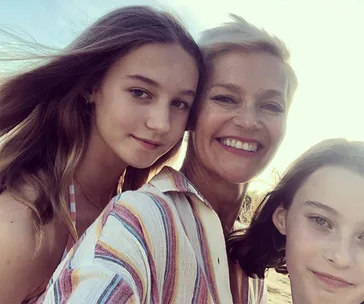 Mother with short blonde hair smiling, taking a selfie with her two daughters on a sunny day.
