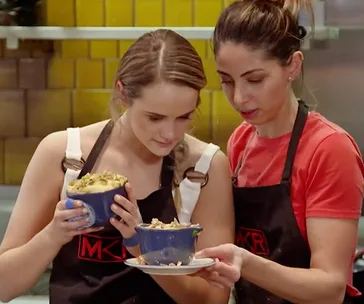 Two women in aprons examining blue ramekins with dishes, inside a kitchen with yellow tiles.