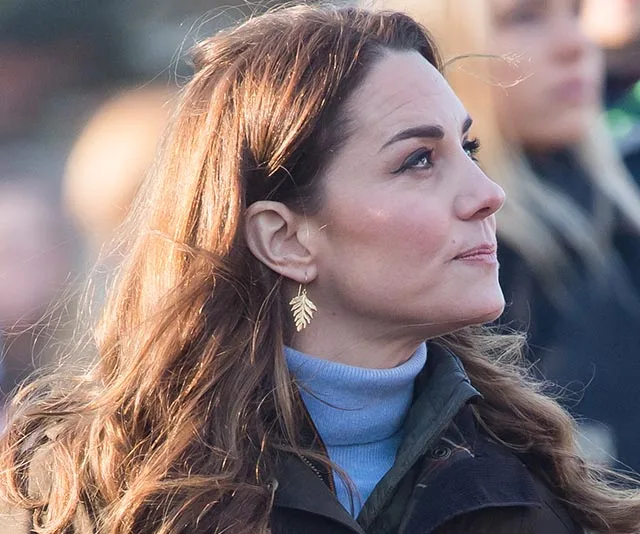 A woman looks upward, wearing a blue turtleneck and gold leaf earrings.