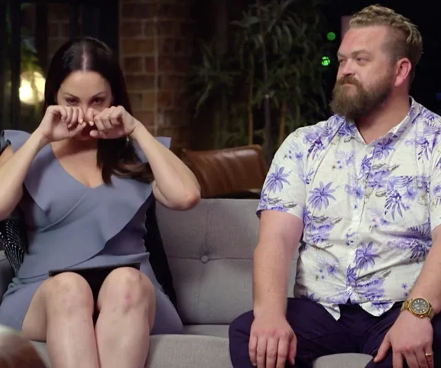 A woman in a blue dress appears emotional, sitting next to a man with a beard in a floral shirt on a couch.