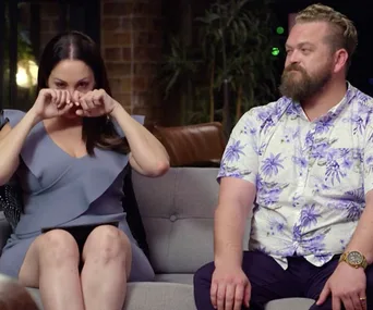 A woman in a blue dress appears emotional, sitting next to a man with a beard in a floral shirt on a couch.
