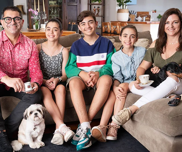 Family of five with two dogs sitting on a couch, smiling together warmly in a cozy living room.