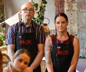 Dan and Steph from "My Kitchen Rules" in aprons, standing together, looking serious.