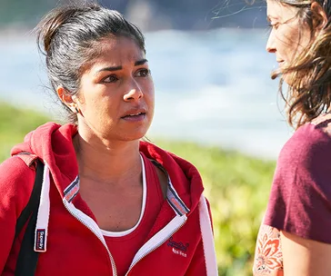 Two women talking outdoors, one in a red hoodie looking serious, ocean and greenery in the background.