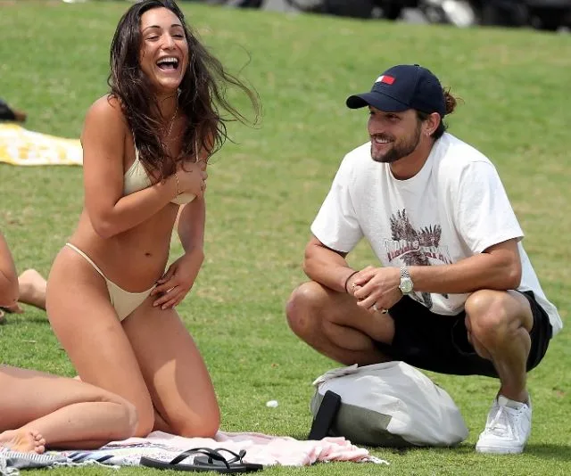 Man and woman smiling and sitting on grass, woman in bikini, man in t-shirt and cap, enjoying a sunny day.