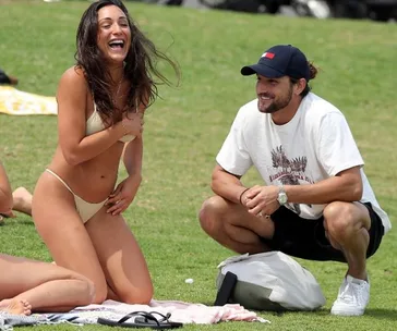 Man and woman smiling and sitting on grass, woman in bikini, man in t-shirt and cap, enjoying a sunny day.