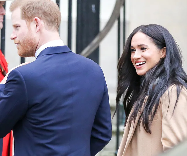 Prince Harry in a blue suit and Meghan Markle in a tan coat smiling outdoors.