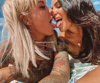 Two women with tattoos playfully touching tongues outdoors under a blue sky.