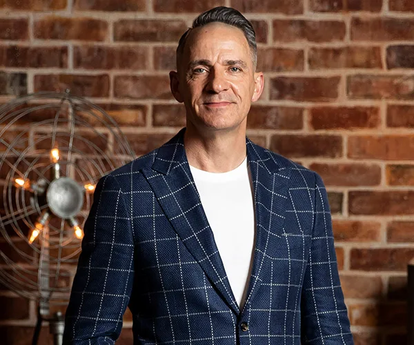 Man in a checkered blazer stands smiling in front of a brick wall, with an industrial-style lamp to the left.