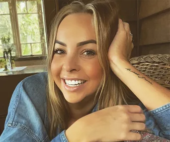 Smiling woman in denim shirt, showing a tattoo on her forearm, indoors with soft lighting and a window in the background.