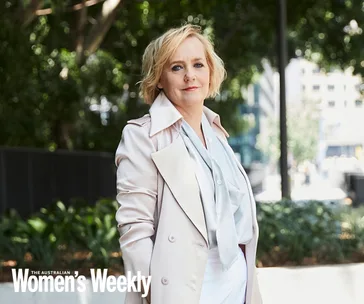 Woman in a light coat stands outdoors, greenery and urban buildings in background; "The Australian Women's Weekly" captioned.