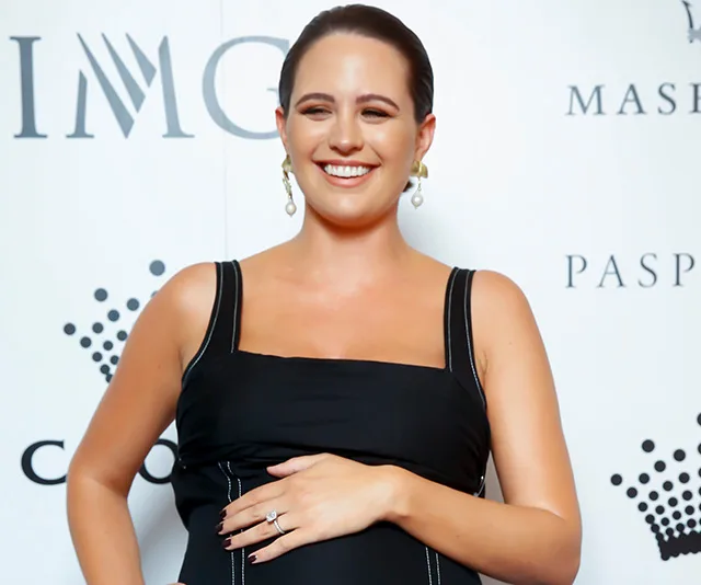 Pregnant woman in a black dress smiling on the red carpet with a hand on her belly.