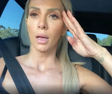 A woman in a car with a seatbelt, one hand raised, looking ahead.
